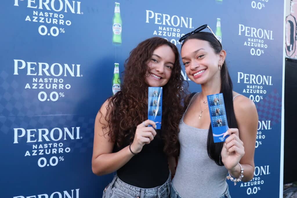 Two women smiling and holding photo booth prints in front of a branded Peroni backdrop featuring the logo and product visuals, illustrating custom branding opportunities for events.