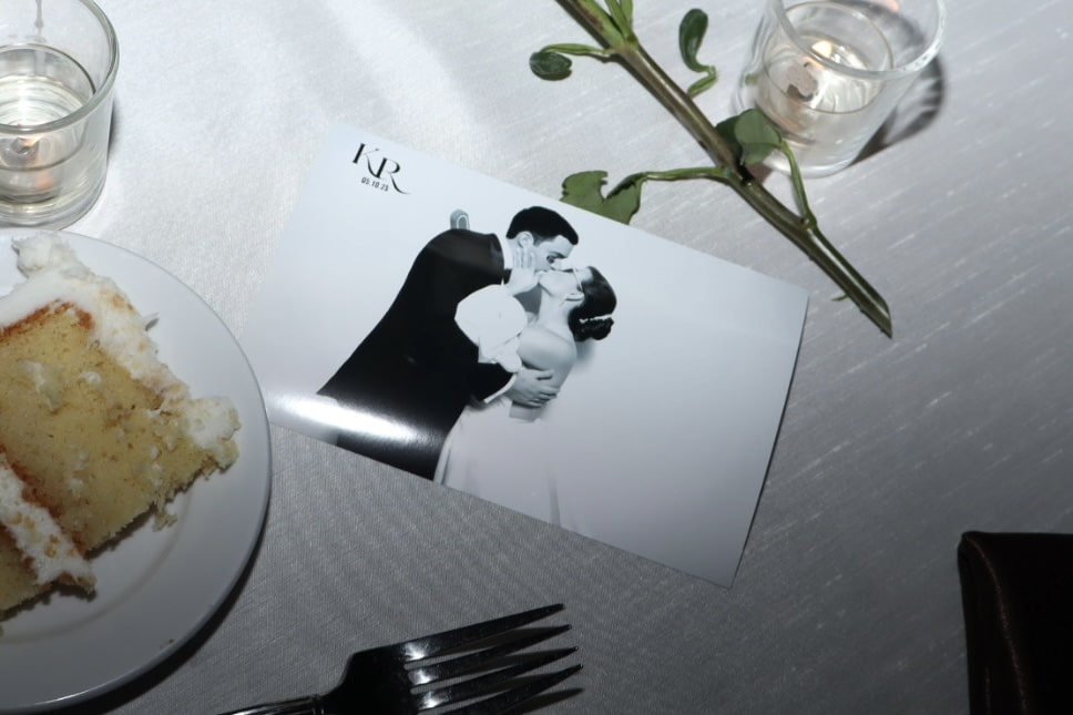 Wedding photo of couple sharing a kiss, surrounded by wedding cake slice, fork, candle, and greenery on a table setting.