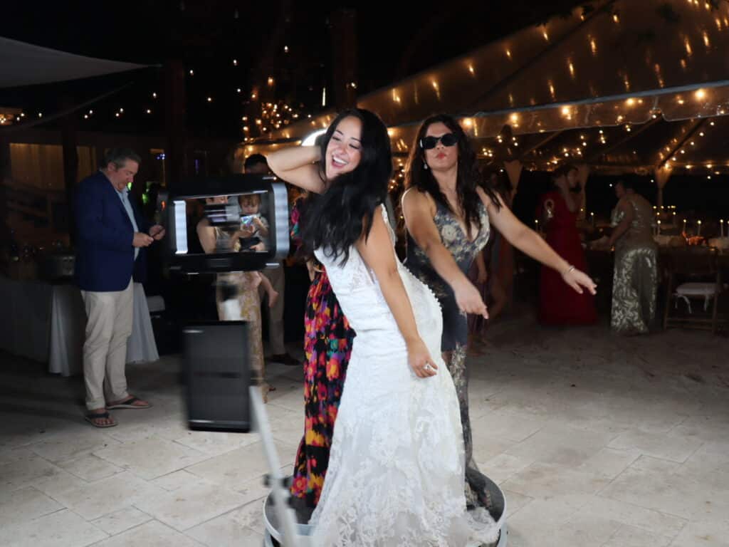 Women posing playfully on a 360 Booth platform, capturing dynamic slow-motion video in a festive event setting with decorative lighting.