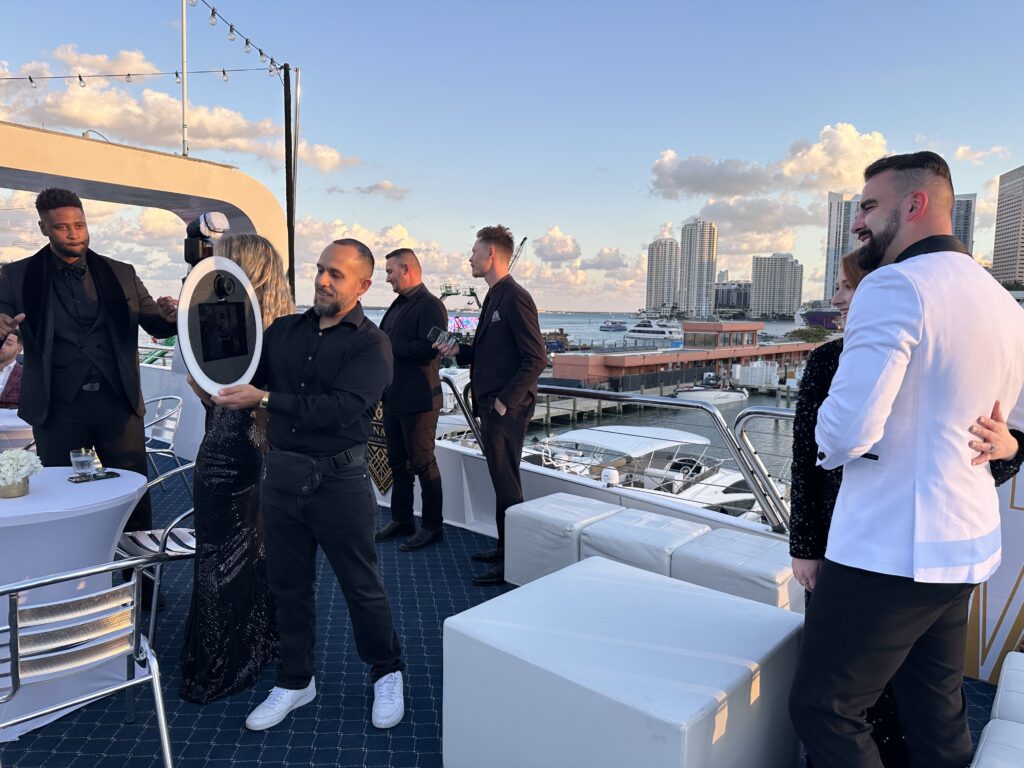 Group of elegantly dressed individuals engaging with a photo booth on a yacht in Miami, showcasing interactive elements for event engagement and social media sharing.