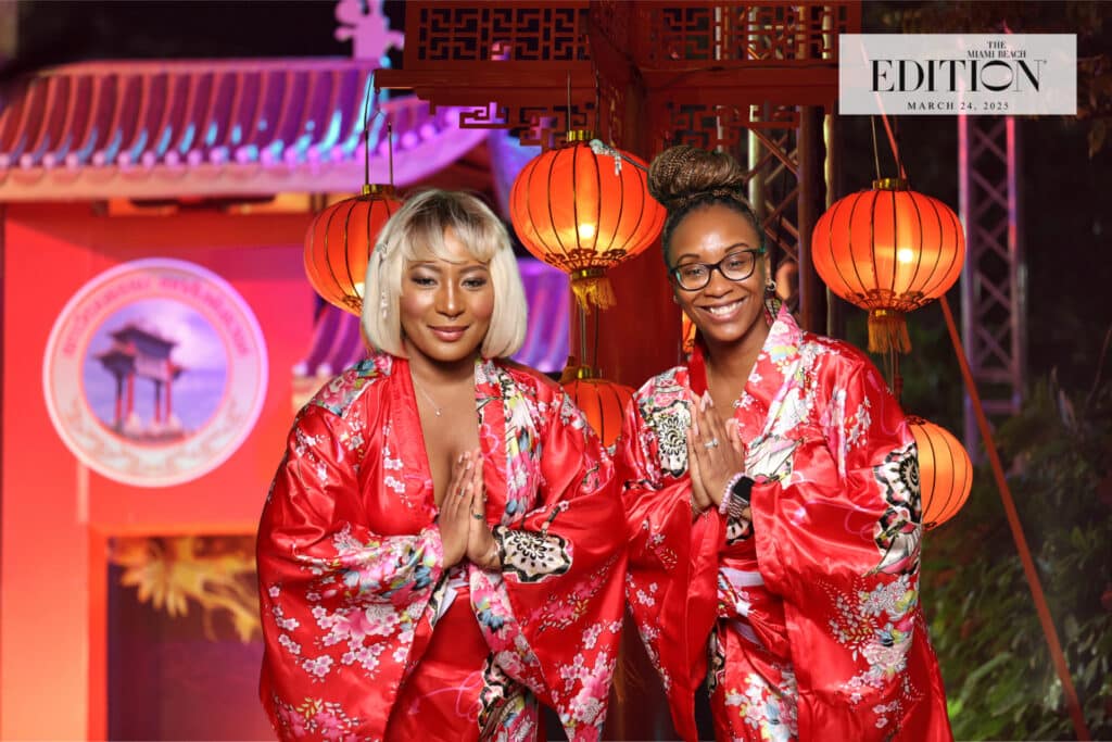 Two women in red kimonos posing with hands together in a gesture of greeting, surrounded by decorative lanterns and a colorful backdrop, representing a vibrant event atmosphere ideal for photo booth sponsorships.