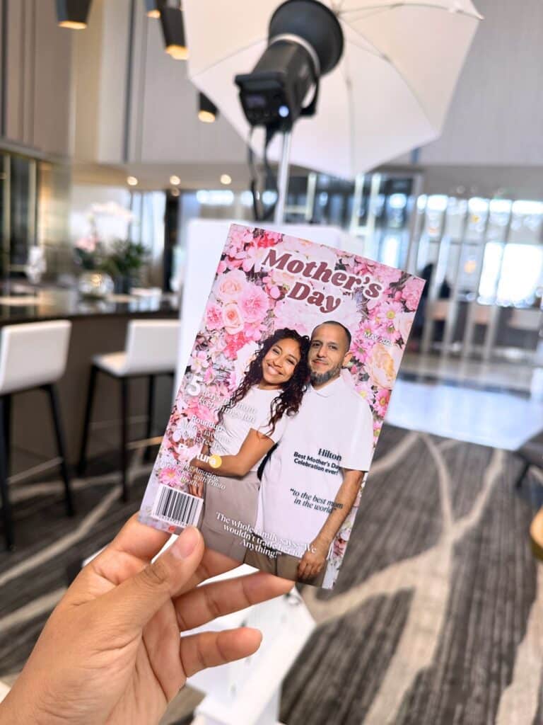 Hand holding a magazine cover featuring a couple celebrating Mother's Day, surrounded by floral designs, with a photo booth setup in the background, highlighting the engagement potential of photo booth activations for brands.